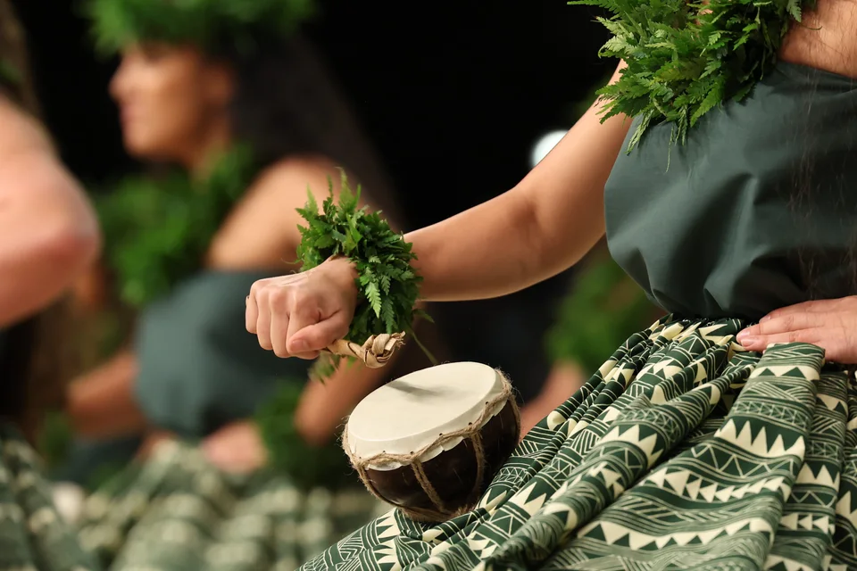 Detail of a dancer holding a hula implement, wearing a green lei at the wrist.