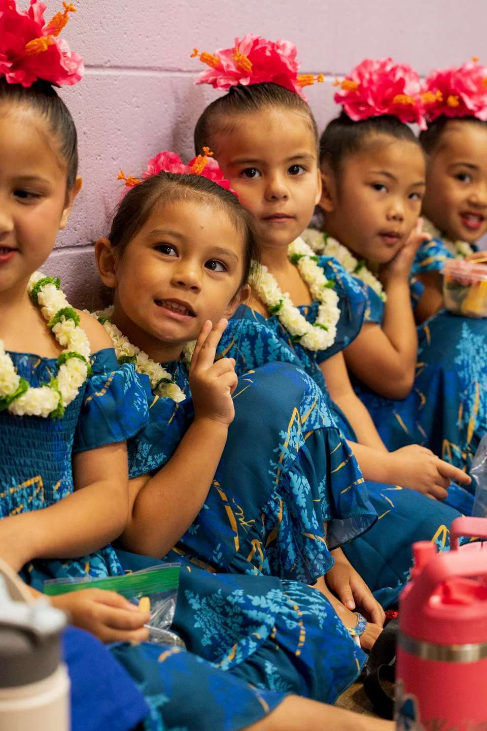 A small group of keiki dressed for hula with bright pink flowers in their hair.